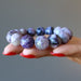 A hand model holds a purple Charoite gemstone bracelet which reveals an array of violet tones and patterns