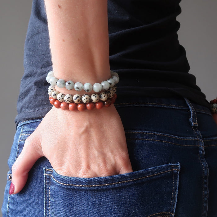 lucia of satin crystals with hands in back jean pockets, wearing set of 3 sesame, red and dalmatian jasper stretch bracelets on each hand.