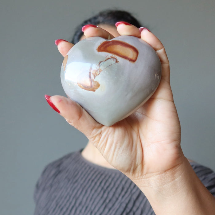 female holding a gray and orange polychrome jasper heart stone in front of her face