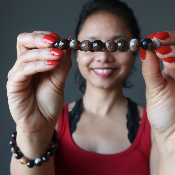 sheila of satin crystals wearing and holding up banded coffee brown and black agate faceted round beaded stretch bracelets