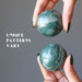 hands holding green and white streaked aventurine spheres to show unique patterns vary in each