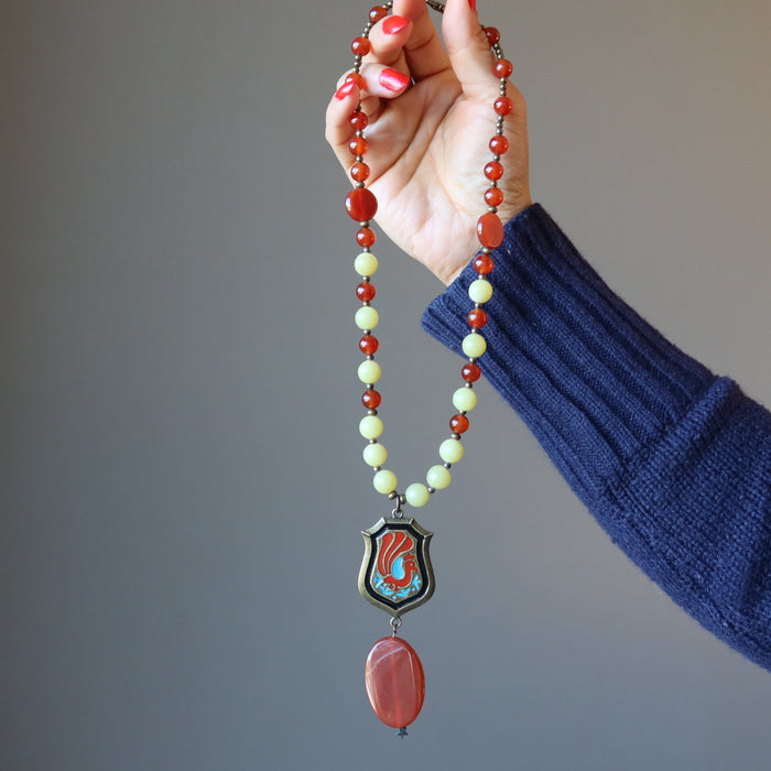 hand holding Necklace made of Carnelian Serpentine and brass beads with oval carnelian hanging underneath painted peacock on brass pendant 