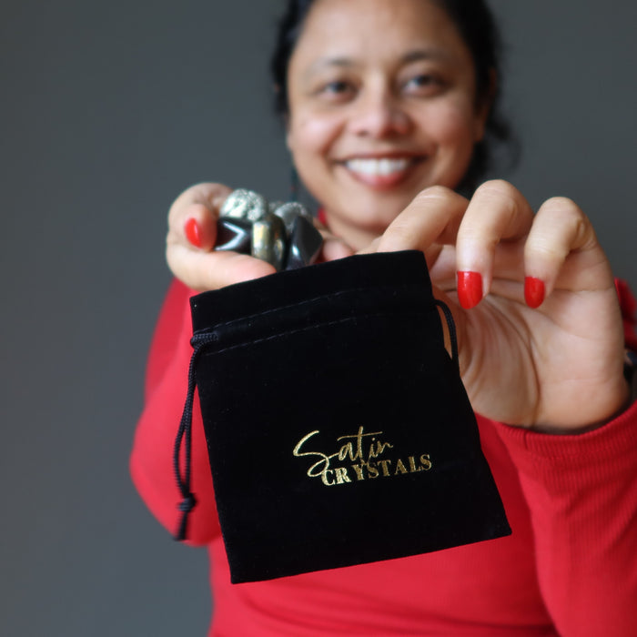 sheila of satin crystals placing hematite, pyrite, chalcopyrite tumbled stones in hand holding black velveteen pouch with gold "satin crystals" print