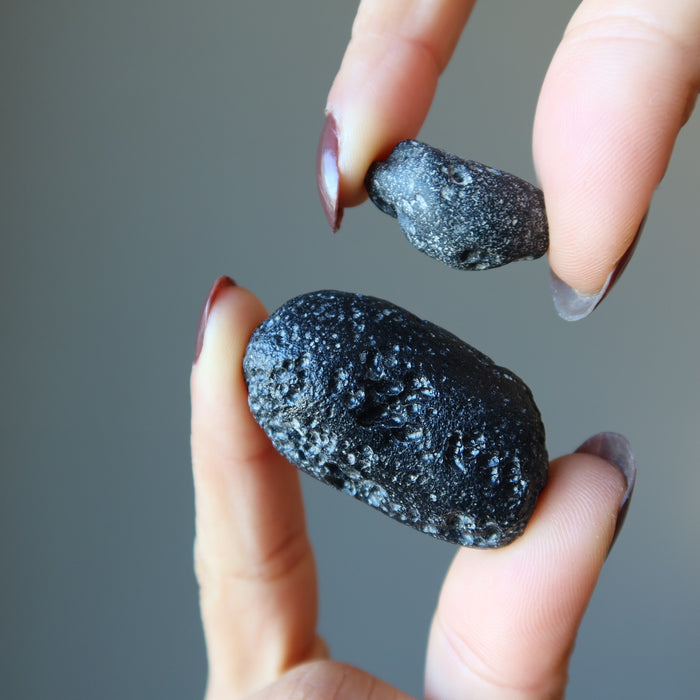 a black tektite held by the tips of fingers from both hands