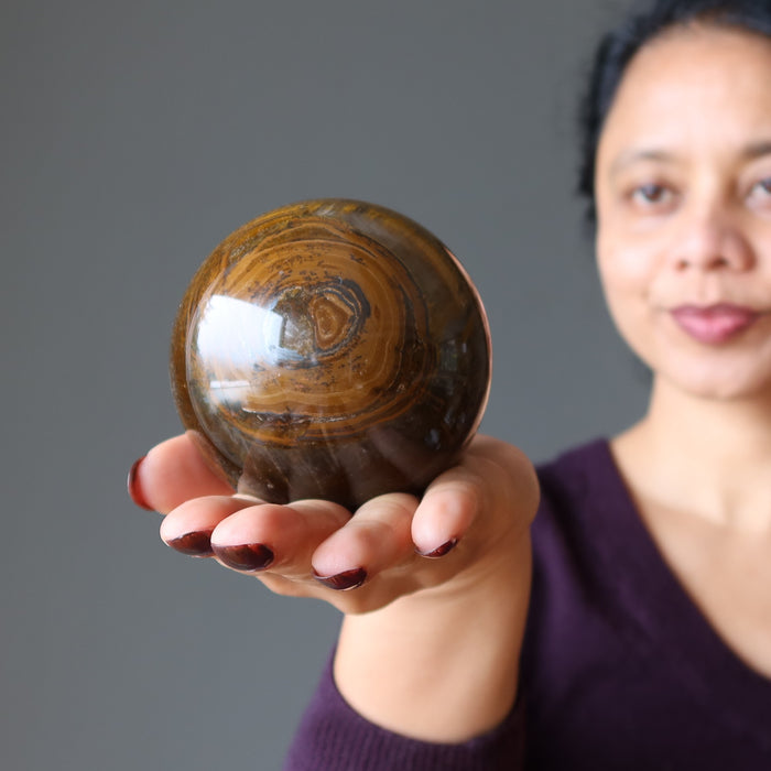 sheila of satin crystals holding a giant brown tiger iron sphere with veins of black hematite